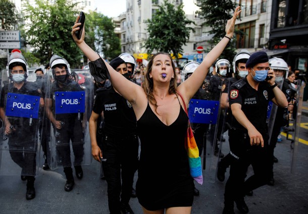 Liana Georgi, eien frau mit langen braunen Haaren, steht auf der straße vor einer Vielzahl an Polizeibeamten und hat eine Pfeife im Mund und streckt ihre Arme in die Höhe.